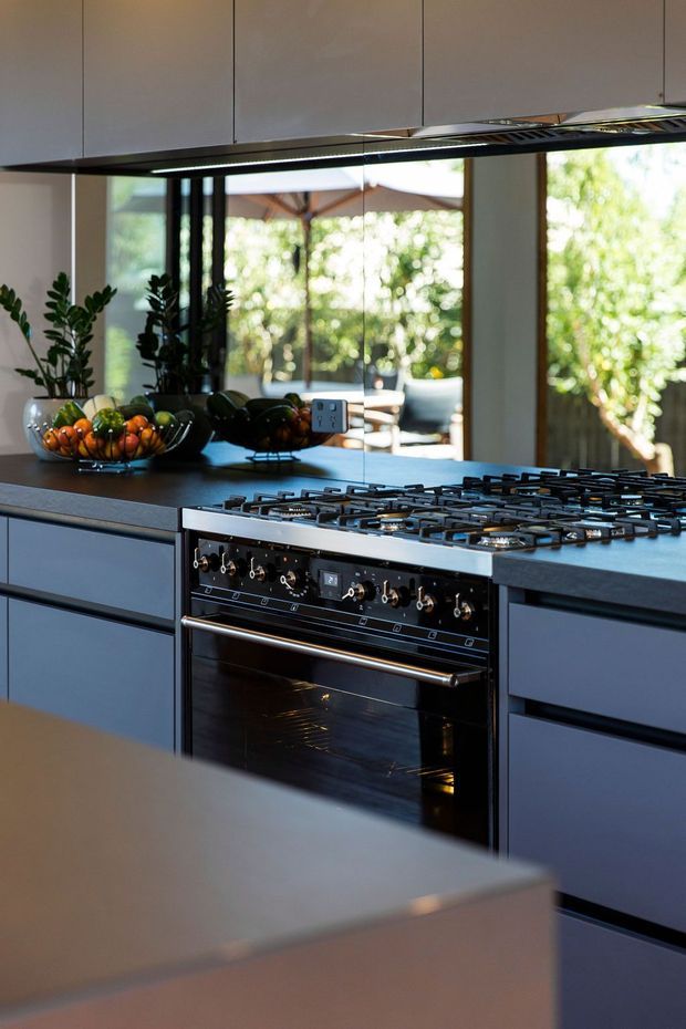A contemporary kitchen viewed close to a large freestanding gas range and oven. Dark blue-grey flat-panel cabinetry runs beneath a dark benchtop, with matching overhead cabinetry and a slim integrated rangehood. A broad opening or glazing behind the kitchen looks onto a bright leafy outdoor area, while bowls of fruit and indoor plants add colour and texture.