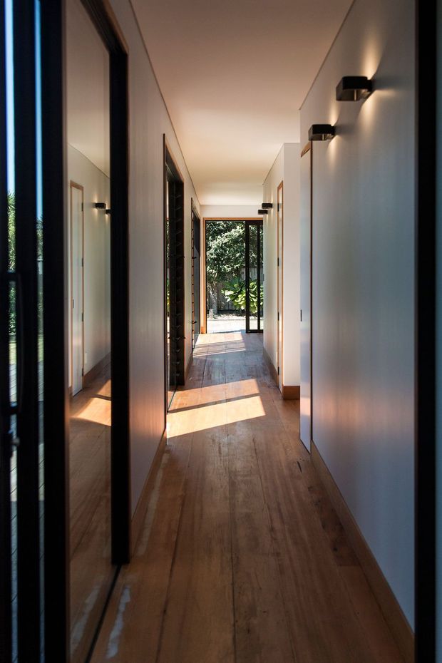 A long, narrow contemporary hallway with warm timber flooring, smooth white walls, black-framed doorways and tall reflective glazing along the left side. Minimal black wall sconces line the corridor, while sunlight enters through the open glazed doorway at the far end and creates bright rectangular patches across the floor. The corridor leads directly toward a leafy outdoor area.