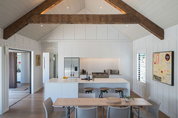 Bright open-plan kitchen and dining area with white vertical wall panelling, a white vaulted ceiling, exposed dark timber trusses, and a large white island with integrated seating. The kitchen includes white cabinetry, stainless-steel appliances, and a light tiled backsplash. A timber dining table with contemporary grey chairs sits in the foreground, with natural light entering through a shuttered window and glazed doorway.