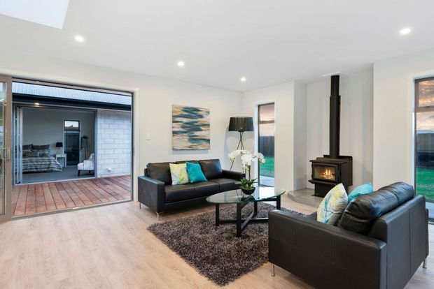 Bright contemporary living room with pale timber flooring, white walls and ceiling, black leather sofas, a glass coffee table, and a dark textured rug. A freestanding wood-burning stove with a tall black flue forms a feature, while large sliding glass doors open to a timber deck and an adjoining covered outdoor area. Recessed ceiling lights and coastal-toned artwork complete the space.