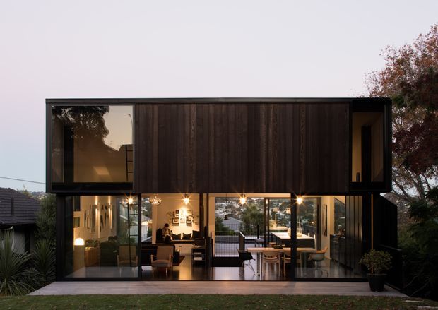 A two-storey contemporary residential house viewed from the rear, with a dark timber-clad upper volume projecting over a fully glazed ground floor. The transparent lower level reveals a warmly lit open-plan living and dining area, while a paved terrace and landscaped garden sit in the foreground. The composition emphasizes strong horizontal lines, black-framed glazing, and a compact modern form.