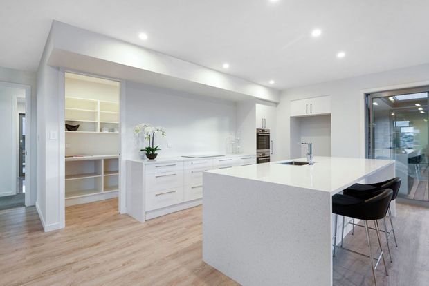 Bright contemporary white kitchen with a large stone island, integrated white cabinetry, built-in shelving, stainless appliances, and two dark bar stools. Light timber flooring, recessed ceiling lights, and glazing to an adjoining outdoor area create an airy open-plan feel.