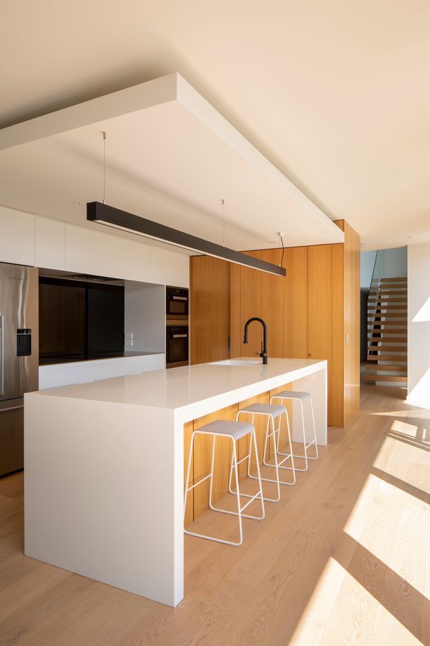 Bright contemporary kitchen with a large white waterfall-edge island, integrated white cabinetry, dark appliances, timber wall cabinetry, and four white bar stools. A linear black pendant light hangs above the island, while an open-riser timber staircase is visible at the rear. Pale timber flooring and strong natural sunlight create an airy, warm interior.