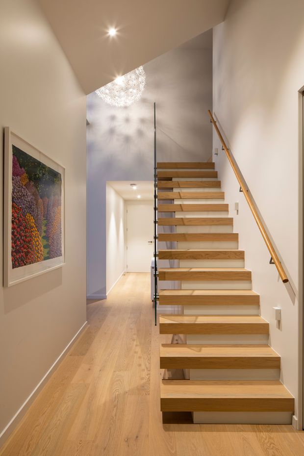 Interior view of a bright residential stairway and hallway. A floating timber staircase with open risers, glass balustrade, and a timber handrail rises beside pale timber flooring and warm off-white walls. Recessed ceiling lights and a decorative pendant cast patterned shadows, while framed artwork is mounted on the left wall and a corridor extends beneath and beyond the stairs.