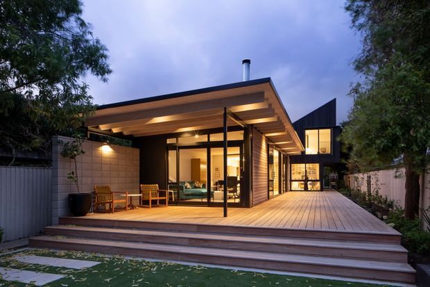 Contemporary single-storey pavilion-style home viewed from the landscaped rear, with a broad low-pitched roof, warm timber soffits and horizontal timber cladding. Floor-to-ceiling black-framed glazing opens onto a generous timber deck with multiple steps, outdoor seating and a covered entertaining area. A taller dark-clad wing is visible behind, with warm interior lighting creating an inviting evening atmosphere.