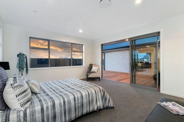 Bright contemporary bedroom with a low-profile bed dressed in a black, white and grey patterned bedspread, a grey carpeted floor, white walls and ceiling, and a compact upholstered armchair. Large windows and sliding glass doors open to a timber-decked outdoor area, with a neighbouring wall and sunset visible beyond.