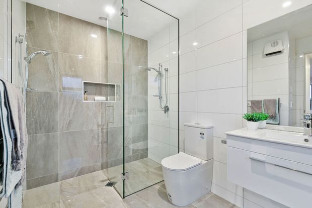 Bright contemporary bathroom featuring a frameless glass walk-in shower with large grey stone-look tiles, white tiled walls, a wall-mounted vanity with integrated basin, and a modern toilet. A recessed shower niche and chrome fixtures provide functional detailing.
