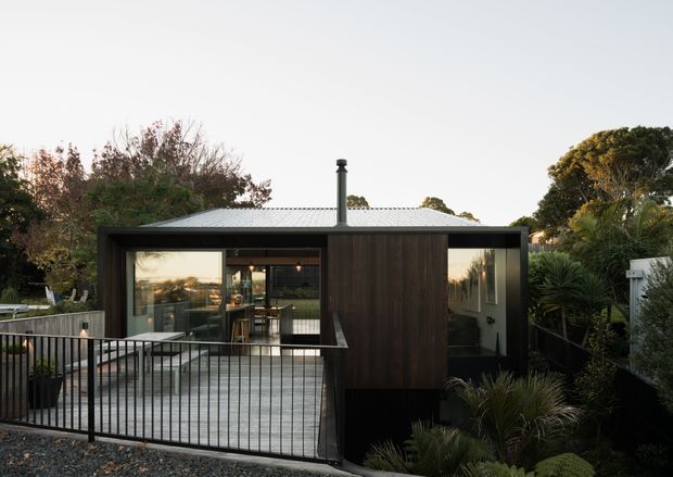 Exterior view of a compact contemporary single-storey home or pavilion, featuring dark vertical timber cladding, large glazed openings, a low corrugated metal roof with a central flue, and a raised deck enclosed by a black metal balustrade. The building is surrounded by dense landscaped planting and mature trees.