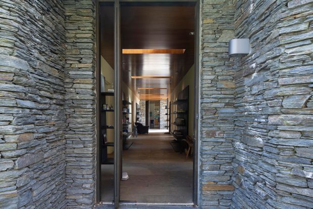 A narrow architectural entry passage framed by substantial stacked-stone walls, with a full-height dark-framed glazed door opening into a long interior corridor. The corridor features a warm timber ceiling, repeated linear ceiling lights or beams, built-in shelving, timber flooring, and a view toward furnished living spaces beyond.