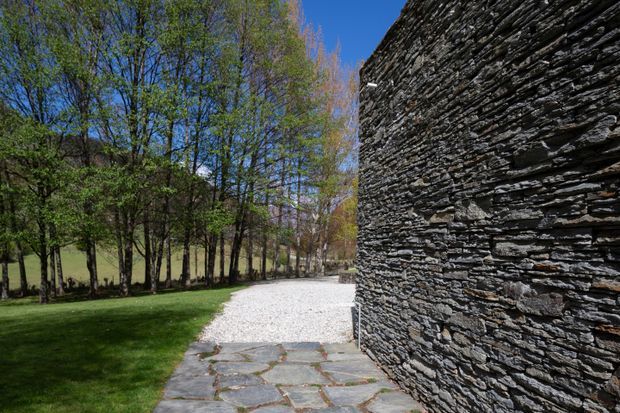 A landscaped outdoor pathway beside a striking dark stacked-stone wall. Large irregular stone paving leads into a pale gravel path, bordered by mature deciduous trees and manicured grass under a clear blue sky.