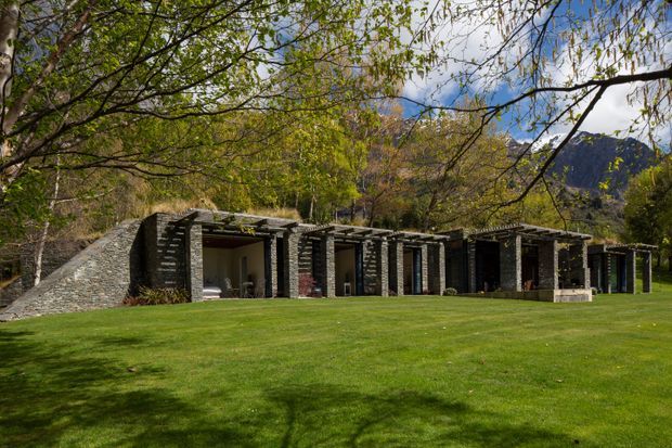Low, elongated stone-clad building set into a landscaped lawn, with a repeating sequence of open bays and dark timber or metal roof structures. Mature deciduous trees frame the composition, with rugged mountains visible in the background. The architecture has a strongly horizontal, site-integrated character and appears to be a pavilion or retreat viewed from outdoors.