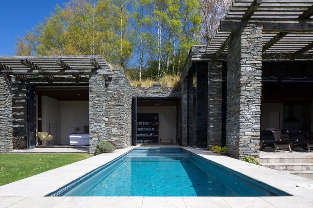 A contemporary luxury residence arranged around a narrow rectangular swimming pool. The facade combines extensive rough stacked-stone walls with dark timber pergolas and large glazed openings, while a manicured lawn, pale paving, and stepped terrace frame the courtyard-like outdoor space. Interior rooms and lounge furniture are visible beneath the covered pergolas, with mature trees and a clear blue sky beyond.