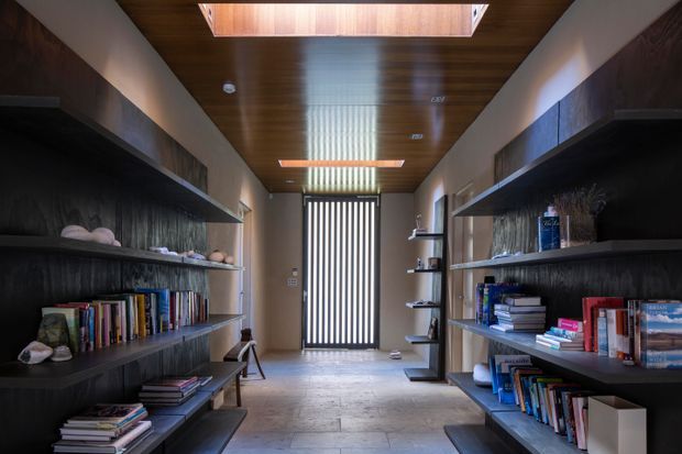 A narrow, symmetrical interior corridor or library lined with dark built-in open shelving filled with books and decorative objects. The space has a warm timber-lined ceiling with skylights, textured dark wall finishes, pale large-format floor tiles, and a vertically slatted glazed door or screen at the far end.
