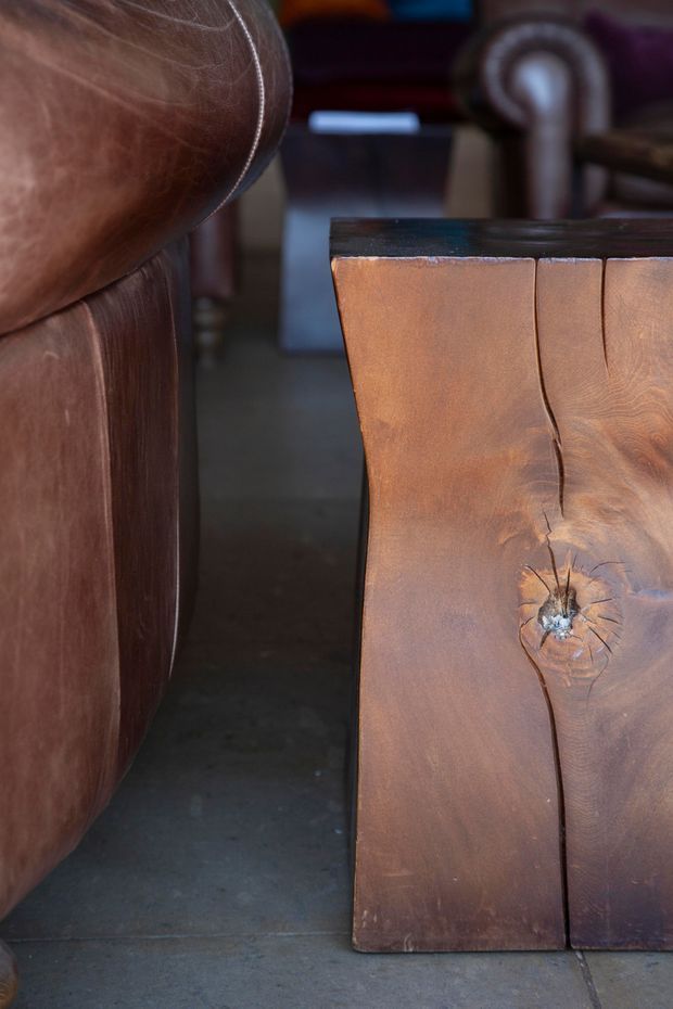 Close-up interior detail of a rustic solid timber side table or console with a live-edge character, prominent natural knots and grain, positioned beside a brown leather armchair on a dark concrete floor. The image emphasizes handcrafted woodwork and warm, tactile materials.