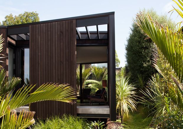 Exterior view of a compact modern pavilion or home extension nestled in dense landscaping. The building has dark vertical timber cladding, black-framed floor-to-ceiling glazing, a projecting flat roof, and clerestory-style upper windows. Lush palms and ornamental grasses surround the structure, with a glimpse of an interior seating area through the glazing.