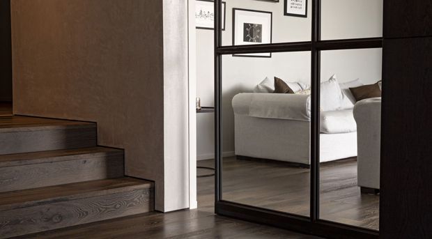 Interior view of a refined residential hallway or stair landing with dark timber stairs and flooring, a white plaster wall, and a large black-framed mirrored sliding door or partition. The mirror reflects a light upholstered sofa, framed wall art, and part of the adjoining living area.