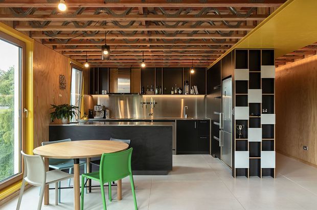 A contemporary open-plan kitchen and dining area with a compact round timber dining table and colourful chairs in the foreground. The kitchen features a dark island and cabinetry, stainless-steel splashback, integrated appliances, and warm under-cabinet lighting. Timber wall lining and an exposed timber-framed ceiling with visible services add strong architectural character, while a black-and-white open shelving partition separates the kitchen from the adjoining space.