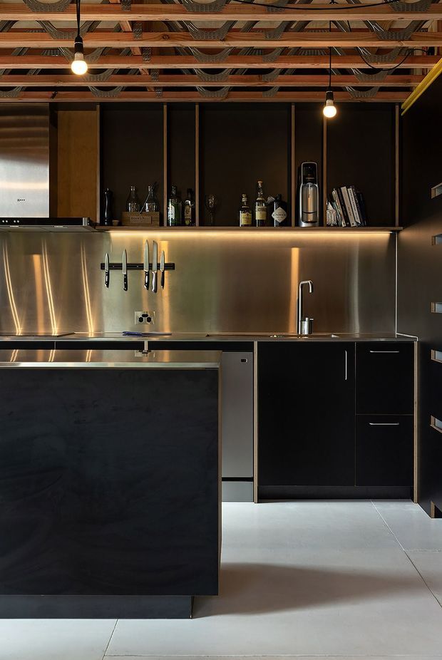 A compact, sophisticated kitchen with black cabinetry, stainless-steel splashback and benchtops, integrated appliances, a large dark island, open shelving with bottles and glassware, and a wall-mounted knife rack. The ceiling features exposed timber beams with decorative metal mesh elements, while warm pendant bulbs provide contrast against the dark finishes.