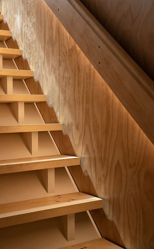 Close-up view of a compact timber staircase with open risers, exposed supporting brackets, plywood side walls, and a substantial timber handrail. The warm natural wood surfaces dominate the narrow stairwell, highlighting the joinery and construction details.