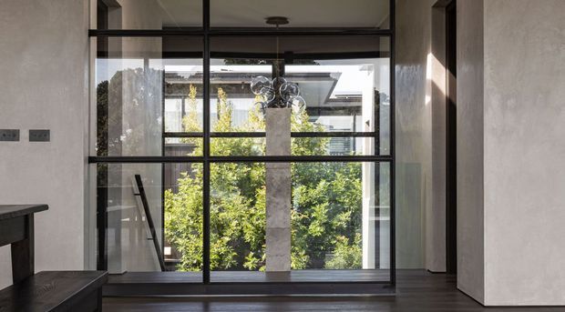 A refined interior view centred on a large black-framed glazed wall or internal screen, with a garden courtyard visible beyond. The space has dark timber flooring, pale textured plaster walls, strong vertical and horizontal framing, and a sculptural pendant light. A partial dark timber bench or console is visible at left, creating a calm, contemporary architectural composition.