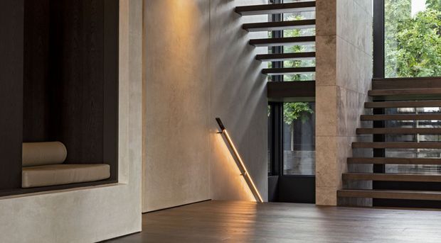 A dramatic contemporary interior entry or stair hall with dark timber flooring, textured pale concrete or plaster walls, a recessed dark timber niche with bench seating, and an open-riser timber staircase. Linear ceiling openings and a slim wall-mounted light create strong bands of natural and artificial light, with greenery visible beyond the glazing.