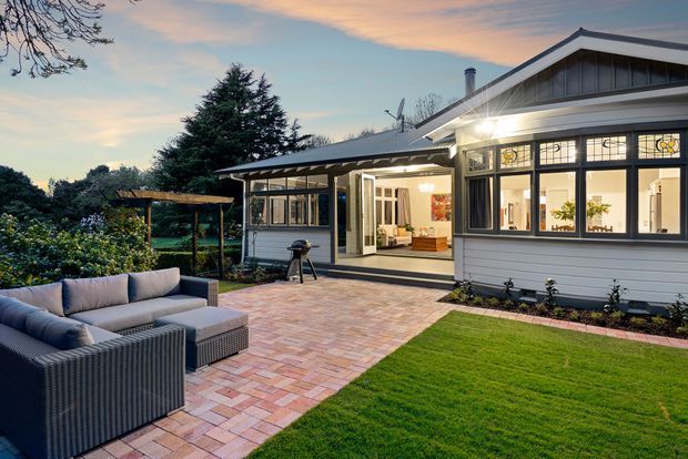 Exterior view of a single-storey suburban home at dusk, featuring a gabled roof, grey horizontal weatherboard cladding, prominent black-framed windows with decorative upper glazing, and an illuminated open-plan interior. A large paved patio extends across the foreground with outdoor lounge seating, barbecue, lawn, garden planting, and a timber pergola.