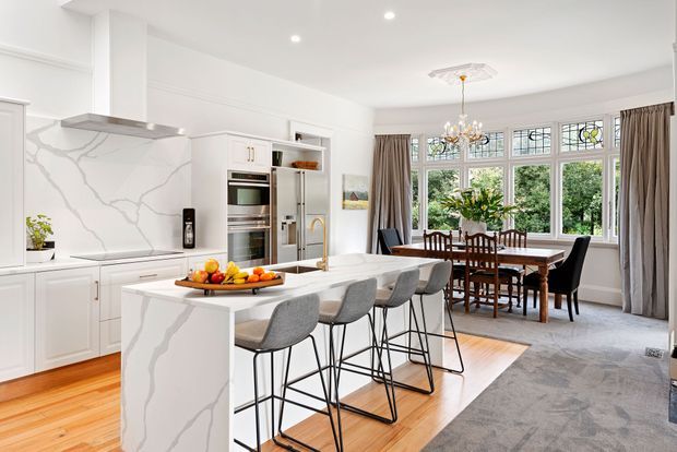 Bright, open-plan kitchen and dining area with white shaker-style cabinetry, a large white marble-look island, integrated stainless-steel appliances, grey upholstered bar stools, and a timber dining table. Decorative leaded or stained-glass-style windows, full-height curtains, timber flooring, and a large area rug create a refined traditional-contemporary interior.