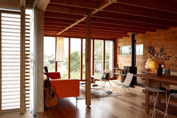 Warm, open-plan timber-lined living and dining area with extensive glazing, exposed timber posts and ceiling battens, polished timber flooring, mid-century furnishings, a red sofa, guitar, lounge chairs, dining table and a freestanding black wood heater. The space has a strong connection to the landscaped exterior through large windows and glazed doors.