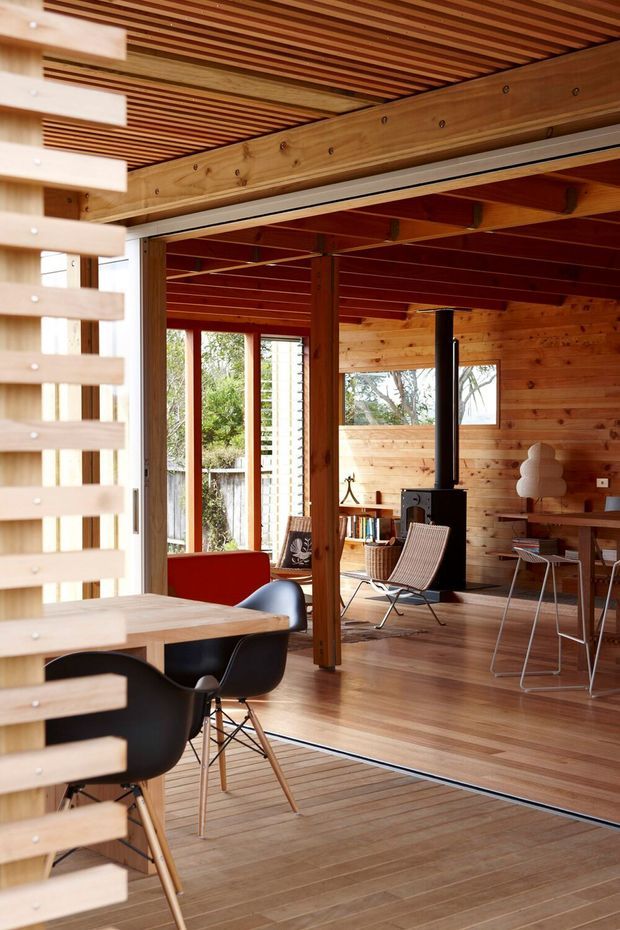 Open-plan timber interior with a dining area in the foreground and a relaxed living space beyond. The room features extensive warm timber wall cladding, timber flooring, exposed beams, a slatted timber ceiling, large glazed openings, a black freestanding wood-burning stove, and mid-century-style furniture. A vertical timber privacy screen partially frames the left side.