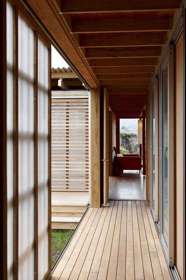 A long, covered timber-lined corridor or gallery with pale timber decking, exposed timber ceiling beams, extensive glazed sliding doors, and translucent screening. The passage connects toward a furnished living area at the far end and opens onto a small planted courtyard or exterior space. The design has a warm, natural, indoor-outdoor character.