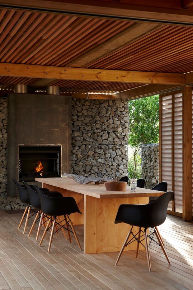 Warm, rustic-modern dining area beneath a slatted timber ceiling. A large timber dining table with black molded chairs sits beside a substantial open fireplace clad in dark metal, with a natural stacked-stone wall behind it. Timber-framed louvred glazing opens to greenery and additional stonework, creating a sheltered indoor-outdoor atmosphere.