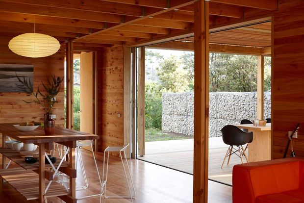 Warm timber-lined open-plan interior with exposed timber beams and posts, polished timber flooring, a dining table with modern chairs, and a compact kitchen or work counter. Large glazed sliding doors open onto a covered outdoor deck, with a gabion stone wall and greenery beyond. A red sofa adds a bold accent in the foreground.