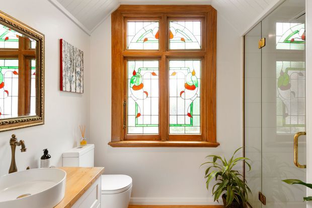 Bright, compact bathroom featuring a timber vanity with a white vessel basin, white toilet, framed mirror, and a glass-enclosed shower. The room is defined by a large stained-glass timber window with colourful botanical-style detailing, white walls and a white sloped-panel ceiling. Brass-toned plumbing and shower hardware, a timber vanity top, and a potted plant add warmth.