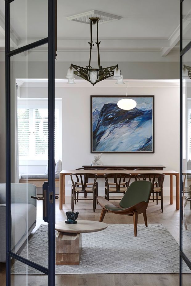 A refined dining and sitting area viewed through open black-framed glazed doors. The room features a timber dining table with sculptural wooden chairs, a green leather lounge chair, a round timber coffee table, a large pale patterned rug, framed abstract artwork, and an ornate hanging chandelier. White walls with substantial decorative cornices and ceiling beams give the space a classic architectural character, while the furniture introduces a mid-century influence.
