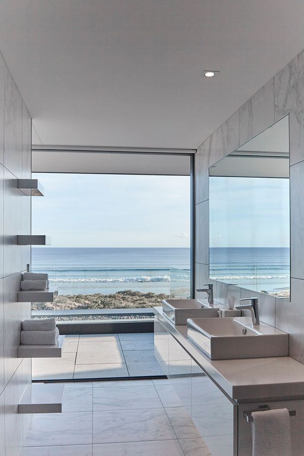 A bright, contemporary bathroom with a double vanity, wall-to-wall mirror, marble-look tiled walls and floor, and a large glazed opening overlooking the ocean and beach. Minimal floating shelving with folded towels provides built-in storage, while recessed ceiling lighting and streamlined fixtures create a clean, refined coastal setting.