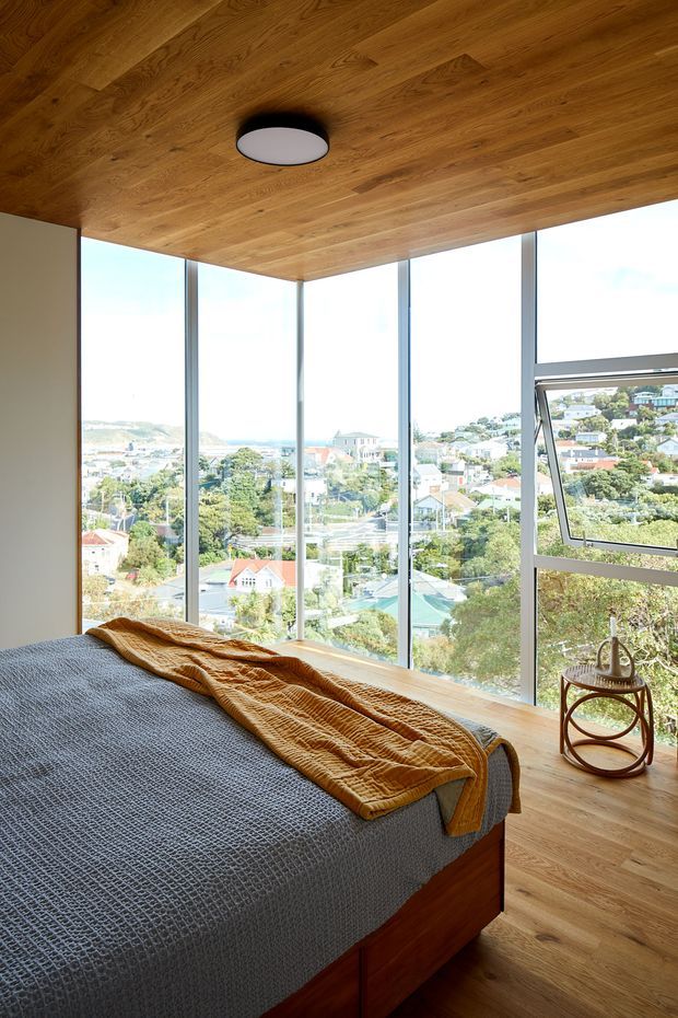 A bright contemporary bedroom with a timber-lined ceiling and matching timber floor, a partially visible timber bed, and expansive floor-to-ceiling corner glazing overlooking a leafy suburban hillside. The room has a simple black-and-white ceiling light, grey bedding, and a mustard textured throw, with a small round side table beside the window.