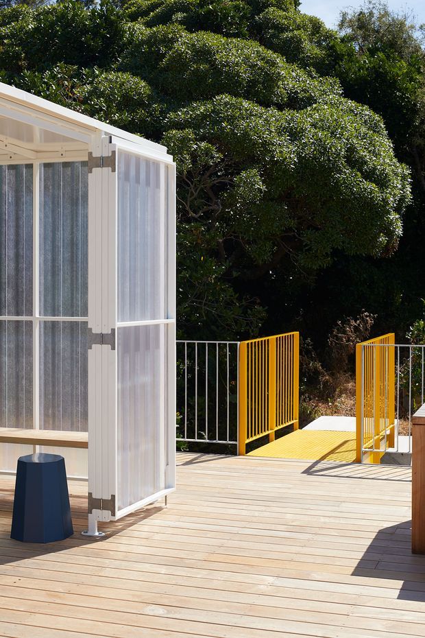 A bright outdoor timber deck beside a lightweight translucent-panel structure or pavilion. The deck has pale horizontal timber boards, white framed folding or hinged screens, and a vivid yellow metal gate and bridge railing leading into dense landscaped greenery. A small dark blue stool sits near the pavilion. The scene is contemporary, sunny, and strongly connected to a garden or elevated outdoor walkway.