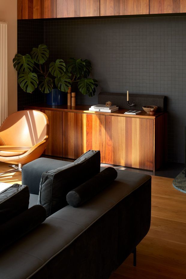 A warm, moody living room featuring a dark upholstered sectional sofa in the foreground, a mid-century-inspired timber sideboard and overhead cabinetry, and a dark small-format tiled feature wall. A tan lounge chair, leafy indoor plant, books, ceramics, and sculptural decor add character. Strong sunlight creates dramatic highlights across the timber joinery and chair, while the timber flooring reinforces the rich, earthy palette.