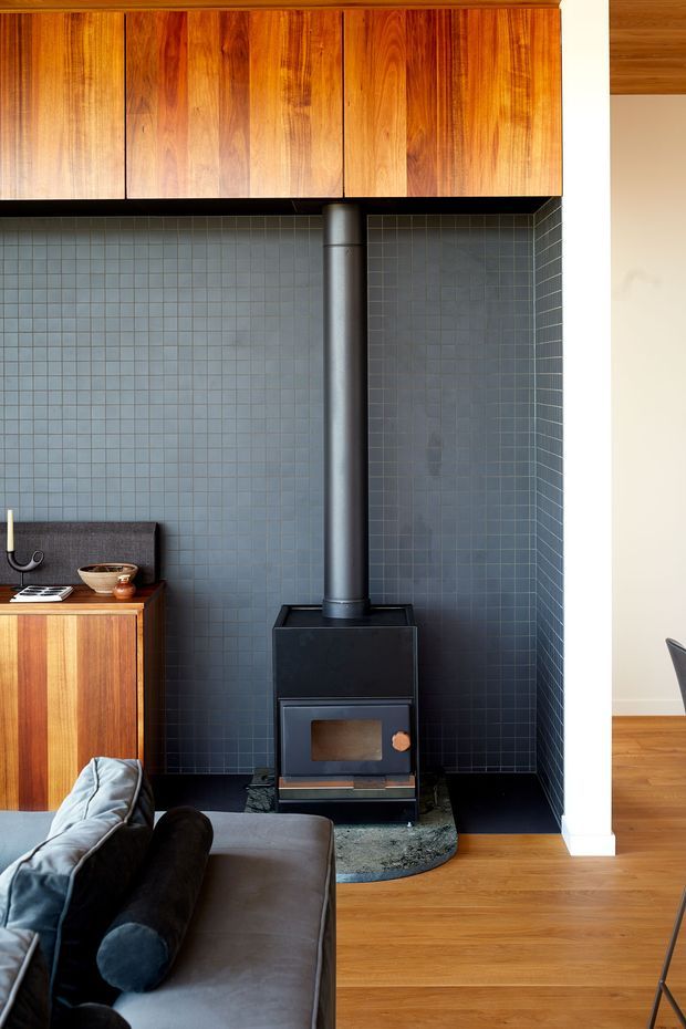 A warm, compact living room focused on a freestanding black wood-burning stove with a tall flue, set against a full-height blue-grey small-format tiled feature wall. Rich timber cabinetry spans the upper wall and continues into a sideboard, while honey-toned timber flooring and a partially visible grey sofa create a mid-century-inspired interior. A white structural wall frames the opening to an adjacent space.