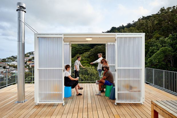 A rooftop or elevated timber deck featuring a compact open-sided shelter with translucent sliding wall panels and a flat roof. Several people gather inside around colorful stools, with a stainless steel flue at the left and safety railings around the deck. Dense trees and suburban houses are visible beyond.