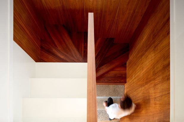 Top-down view of a compact modern stairway with a richly timber-lined ceiling and walls, white plaster walls, and a central timber balustrade or divider. A person is visible descending the stairs, which have speckled grey stone or concrete treads. The strong wood grain and geometric composition create a warm, architectural interior.