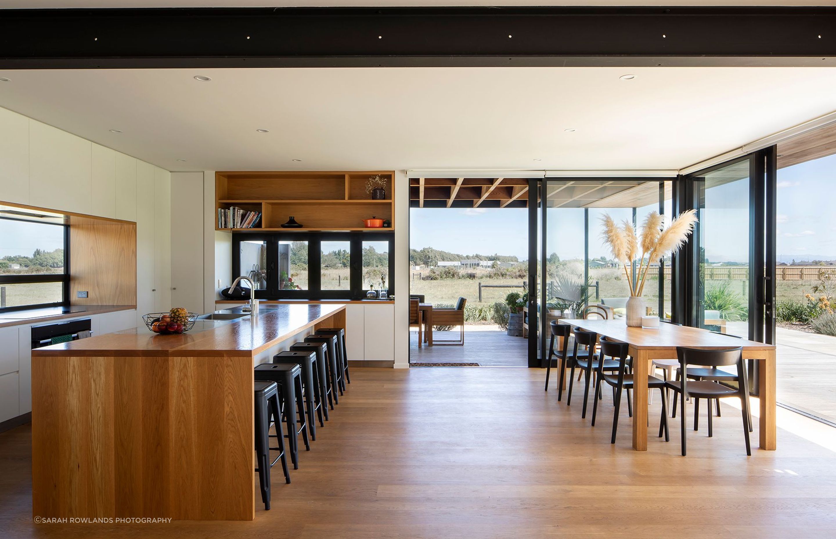 The open-plan kitchen and dining space looking through to the outdoor room and the rural landscape. From the kitchen, the homeowners can also observe visitors coming up the driveway.