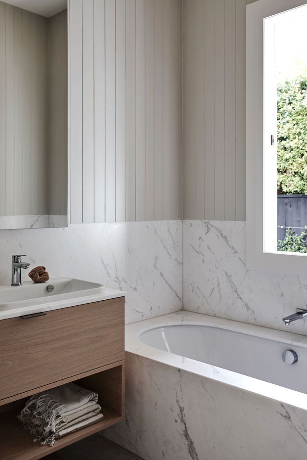 Bright contemporary bathroom featuring a white freestanding-style built-in bathtub, floating timber vanity with integrated basin, large marble-look wall cladding, and vertical white wall panelling. A mirror, chrome tapware, and a large window provide reflective light and views to greenery outside. The palette is light, neutral, and refined.