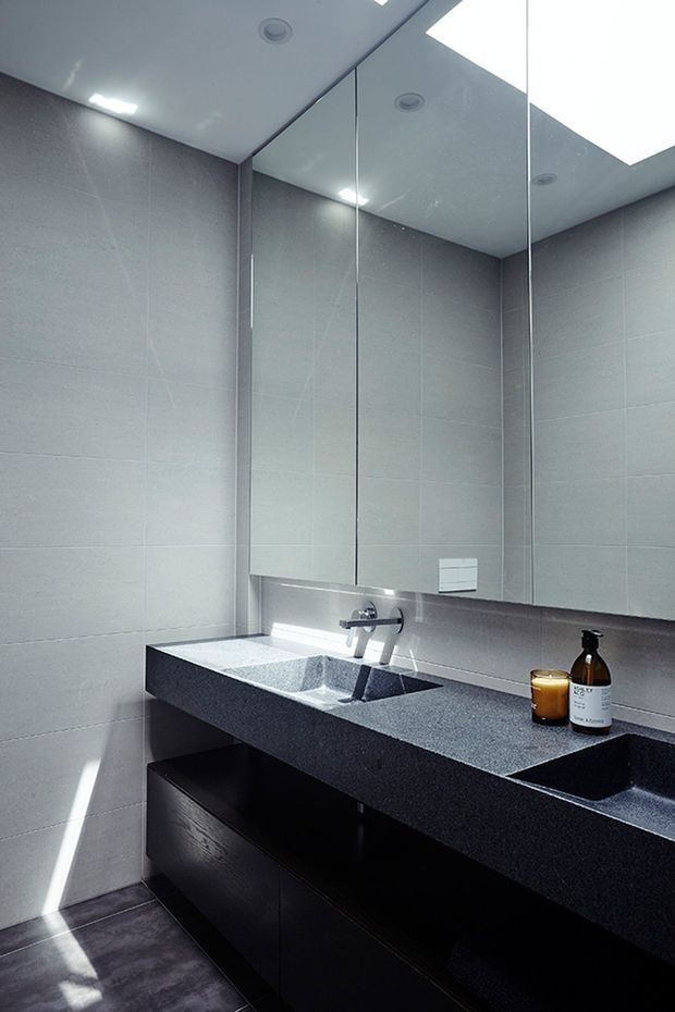 A contemporary bathroom vanity with a long dark stone benchtop, integrated double basins, wall-mounted chrome tapware, dark timber cabinetry, large-format grey wall and floor tiles, and a full-width mirrored wall. Recessed ceiling lighting and a bright skylight-style ceiling panel illuminate the compact space.