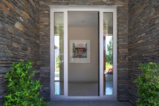 A contemporary entryway framed by dark, horizontally layered stone walls. A glazed grey-framed pivot or hinged front door is flanked by narrow sidelights, opening into a minimalist interior hall with artwork, pale walls, and polished flooring. Green shrubs soften the exterior threshold.