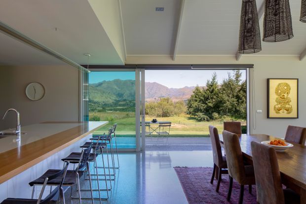 Open-plan kitchen and dining area with a long timber island and bar seating, dark cabinetry, polished light flooring, and a large floor-to-ceiling glazed opening leading to an outdoor terrace and expansive mountain landscape. A dining table with upholstered chairs occupies the foreground, while woven pendant lights and framed artwork add warmth.