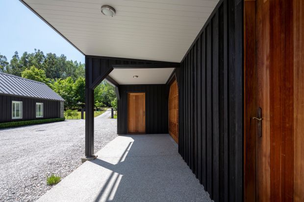 A covered exterior walkway or entry porch beside a dark vertical-clad building. The space features black timber cladding, warm timber doors, a white-lined soffit, concrete paving, and a simple post-supported roof. A matching dark-clad outbuilding and gravel courtyard are visible beyond.