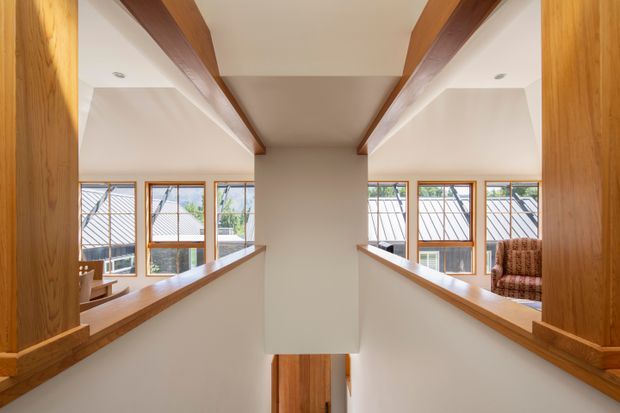 Bright interior view of a split-level landing or hallway with warm timber-framed openings, timber ceiling beams, built-in timber ledges, white walls, and large windows overlooking neighbouring rooftops. A partial seating area is visible beyond the right opening.