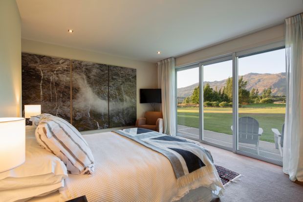 A serene, contemporary bedroom with a large bed dressed in neutral white and beige linens, a dark striped throw, and matching bedside lamps. A dramatic multi-panel nature artwork forms a feature wall behind the bed, while full-height sliding glass doors and sheer curtains open to views of a lawn, trees, and sunlit hills. The room has soft carpet, recessed ceiling lighting, and a warm, relaxed palette.