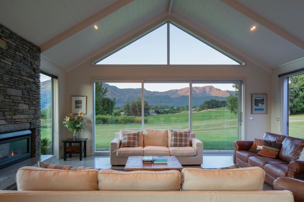Spacious living room with a vaulted, timber-lined ceiling and prominent triangular clerestory window above full-height glazing. A large stone-clad fireplace anchors the left side, while sofas and leather seating face inward around a low coffee table. The glazing overlooks open green fields and mountains beyond.